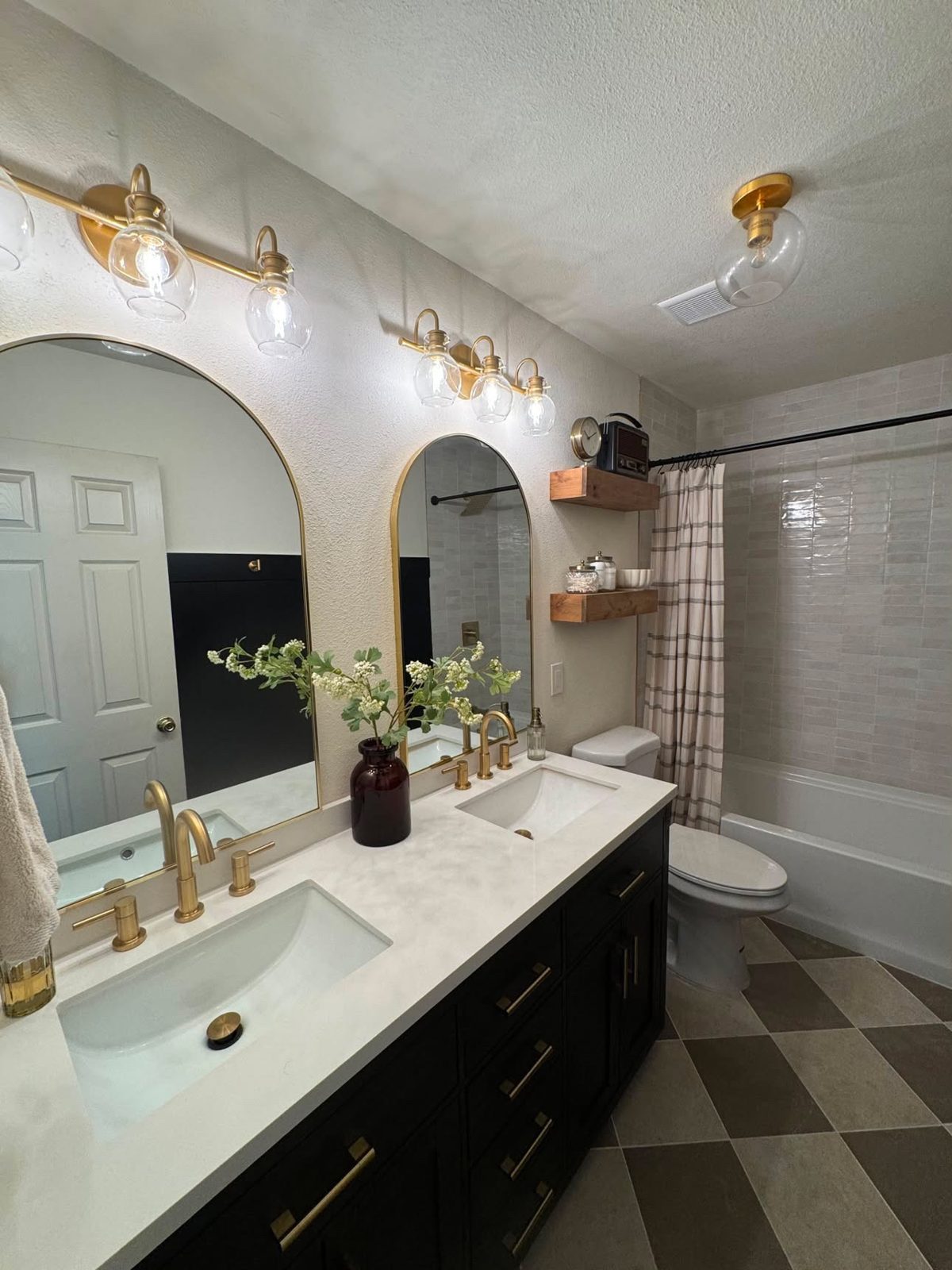 Finished bathroom with a double vanity, black cabinets, arched gold mirrors and a checkered tile floor